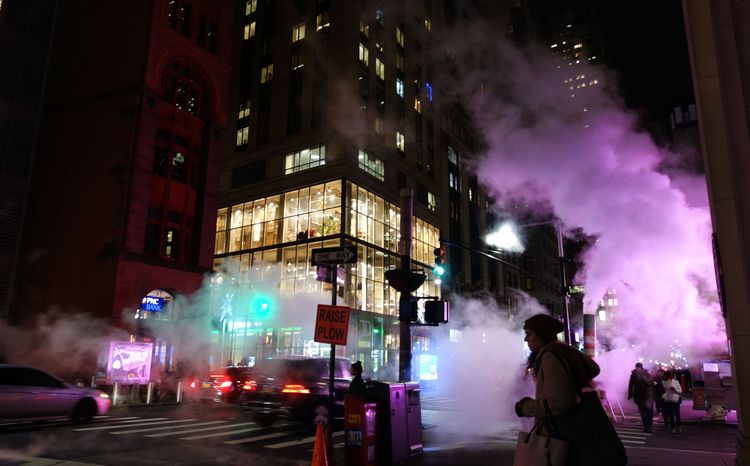 Large clouds of steam billow from nighttime streets. Neon lights lend vivid color to their forms.