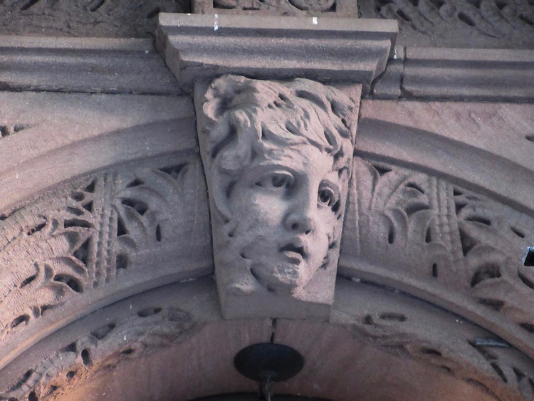 A stone face, looking wind-weary, peers out from the carved keystone of a building façade.