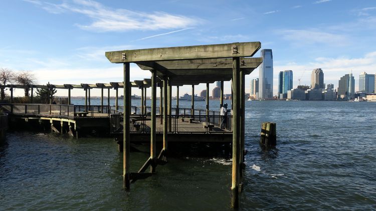 A pier-like structure spirals out into the confluence of river and tide. A person looks out on skyscrapers across the water.